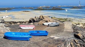 Una playa rocosa con varios barcos, un faro a lo lejos y cielos azules despejados.