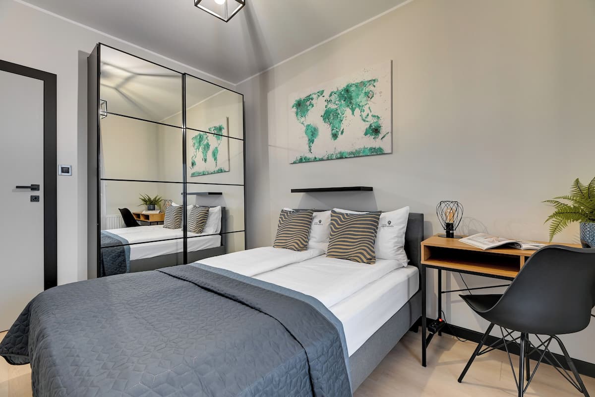 A modern bedroom with a large bed, a desk, and a chair. There is a world map on the wall and a mirror on the door.