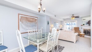 A modern living room with a glass dining table, white chairs, a beige sofa, and a fireplace.