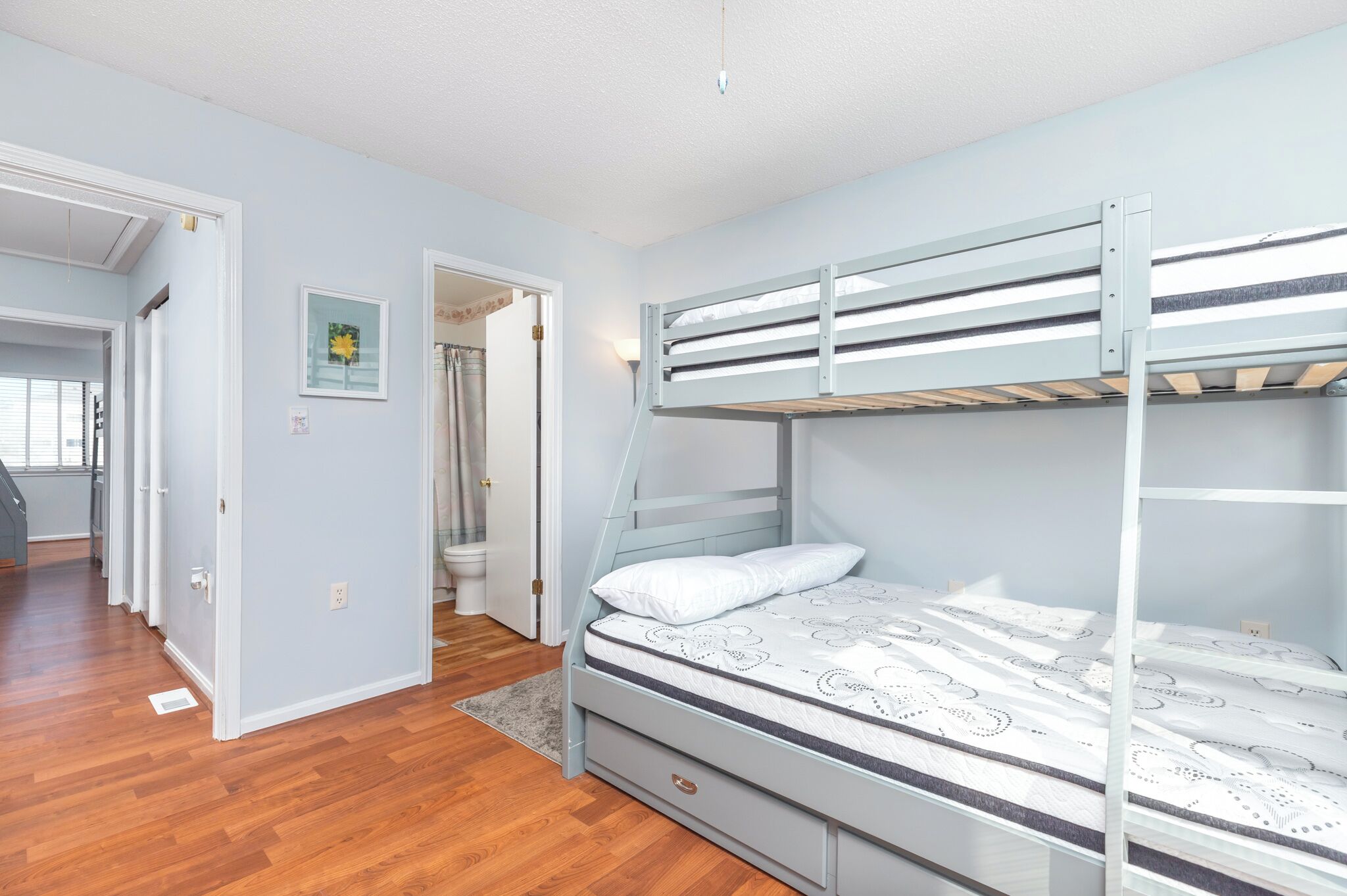Ein Zimmer mit Etagenbett, Holzboden, einer Tür zu einem anderen Raum und einem gerahmten Bild an der Wand.