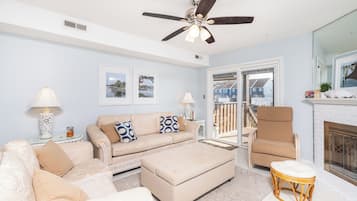 A living room with a beige sofa, a white ottoman, a wicker chair, and a ceiling fan.
