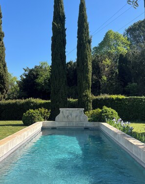 A rectangular pool with a concrete edge, surrounded by well-maintained hedges and tall trees.