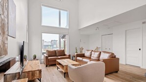 A spacious living room with a large window, a brown leather sofa, a wooden coffee table, and a television.