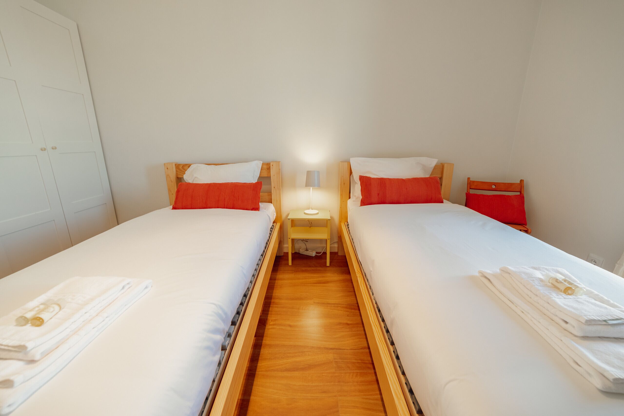 A twin room with two beds, each with white bedding and red pillows, a small bedside table with a lamp, and a wooden floor.