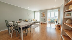 A spacious living area with a dining table, chairs, a sofa, and a coffee table. There is a bookshelf and a window with curtains.