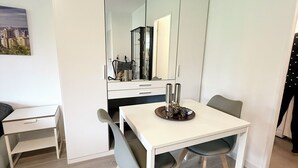 Ein moderner, minimalistischer Essbereich mit einem weißen Tisch, zwei Stühlen und einem dekorativen Mittelstück. Ein großes Spiegelbild zeigt den Raum, und ein kleiner Beistelltisch ist links zu sehen.