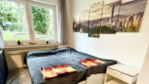Ein Schlafzimmer mit Bett, Nachttisch, Fenster mit Blick auf Grünflächen und einem Stadtbild an der Wand.