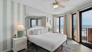 Ein Schlafzimmer mit einem Bett, Nachttischen, einer Lampe, einem Deckenventilator, einem Fenster mit Vorhängen und Blick auf den Strand.