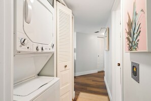 Ein Waschraum mit einer Frontlader-Waschmaschine und einem Trockner, einem Holzboden und einem Gang zu einem Raum mit einer Tür und einer Pflanze.