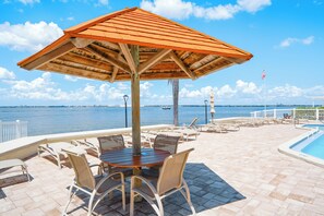 Een zwembadterras met een houten parasol, ligbedden en uitzicht op het water.