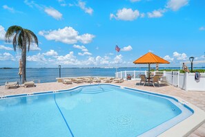 Ein Schwimmbecken mit Liegestühlen und Sonnenschirm, mit Blick auf ein Gewässer und eine Stadtkulisse in der Ferne.