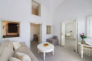 Un soggiorno spazioso con un divano color beige, un tavolino bianco rotondo con della frutta e vista su una cucina attraverso una finestra.
