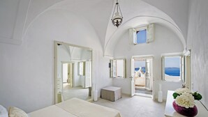 A spacious bedroom with a large bed, a dresser, and a view of the sea through an open door. - Villa Calliope (Santorini)
