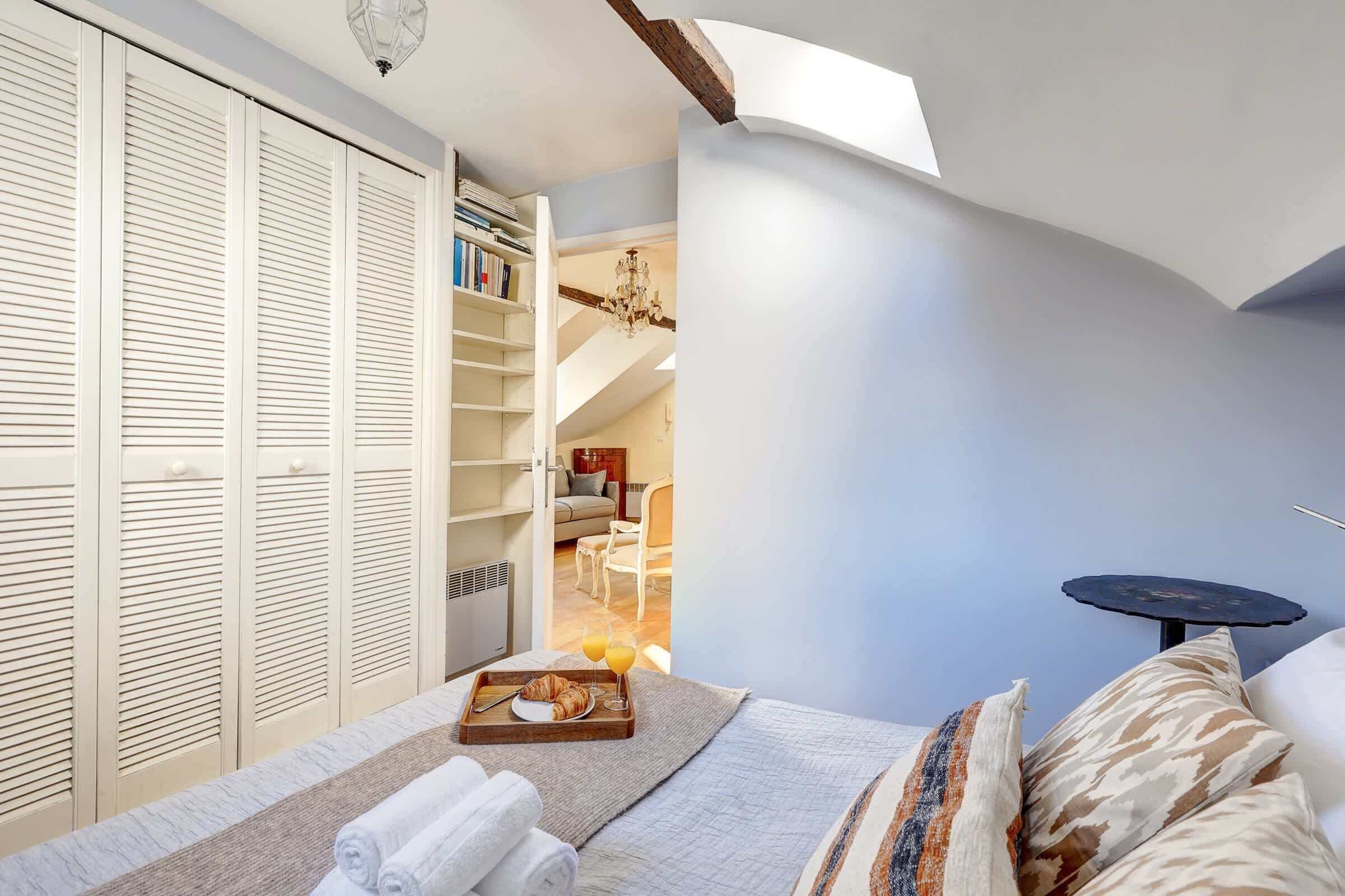 A bedroom with a bed, a bedside table, a tray with croissants and juice, and a view of a dining area.