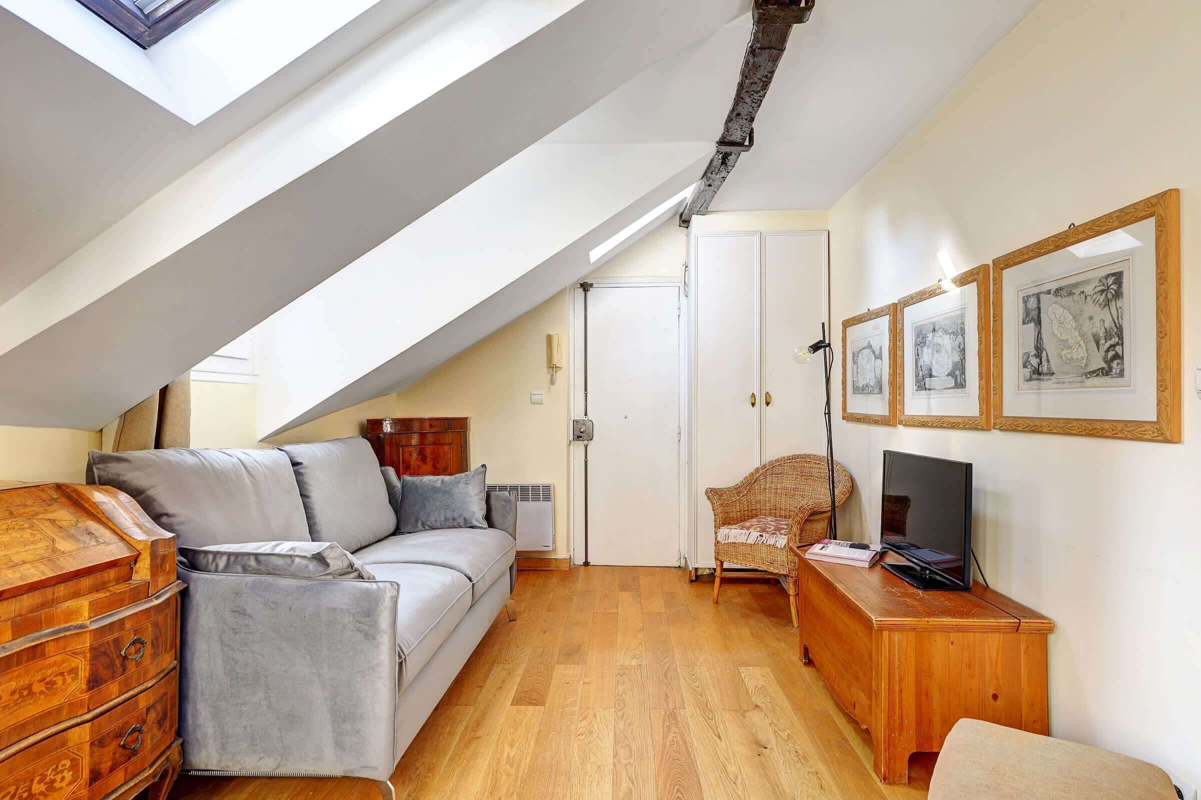 A cozy attic room with a sloped ceiling, a sofa, a wooden dresser, a television, and framed artwork on the wall.