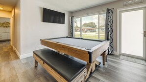 Une table de billard dans une pièce dotée d’une grande fenêtre, d’un plancher en bois et d’un téléviseur à écran plat fixé au mur.