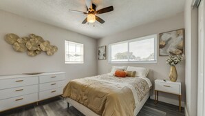 Une chambre à coucher comprenant un lit, une table de chevet, une commode, un ventilateur de plafond et une fenêtre avec des stores.