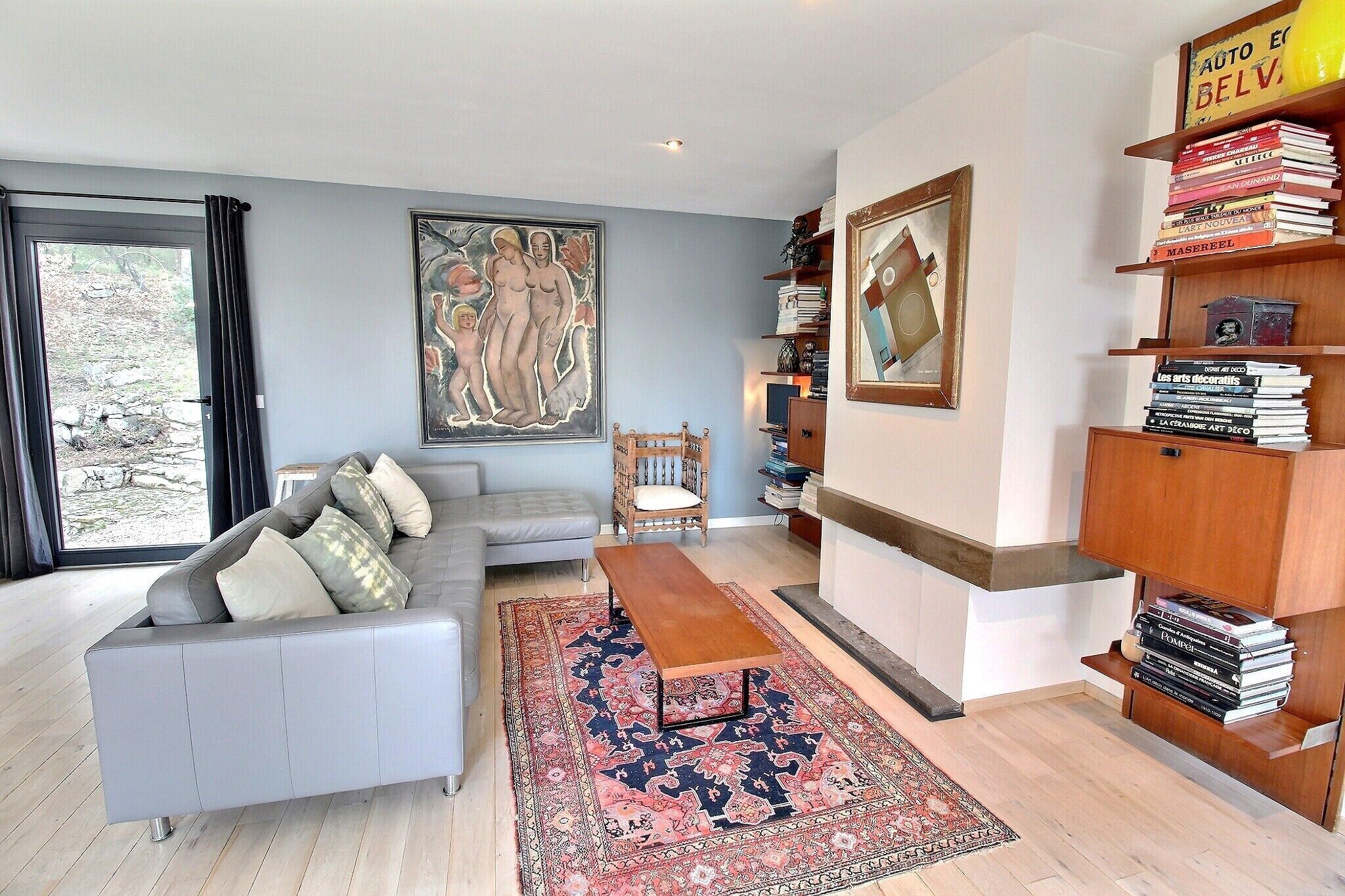 A modern living room with a grey sofa, a wooden coffee table, a large rug, a bookshelf, and a painting on the wall.