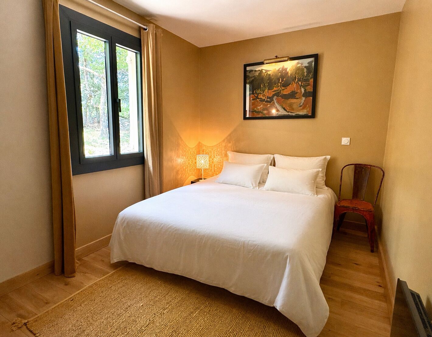 A bedroom with a bed, a nightstand, a lamp, a chair, a painting on the wall, and a window with curtains.