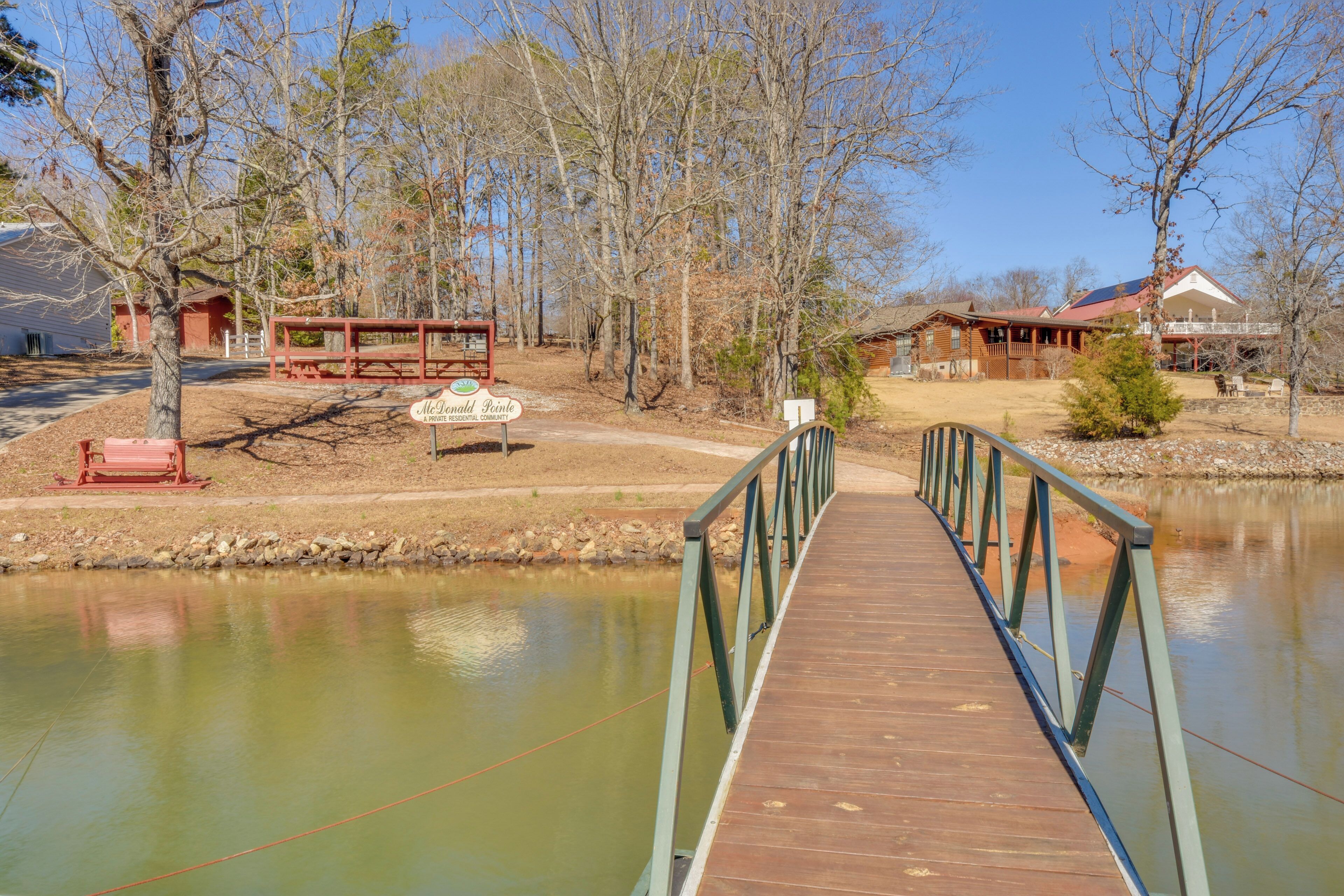 A wooden bridge over a calm body of water, leading to a rustic cabin with a red roof and a sign reading "McDonald Ridge".