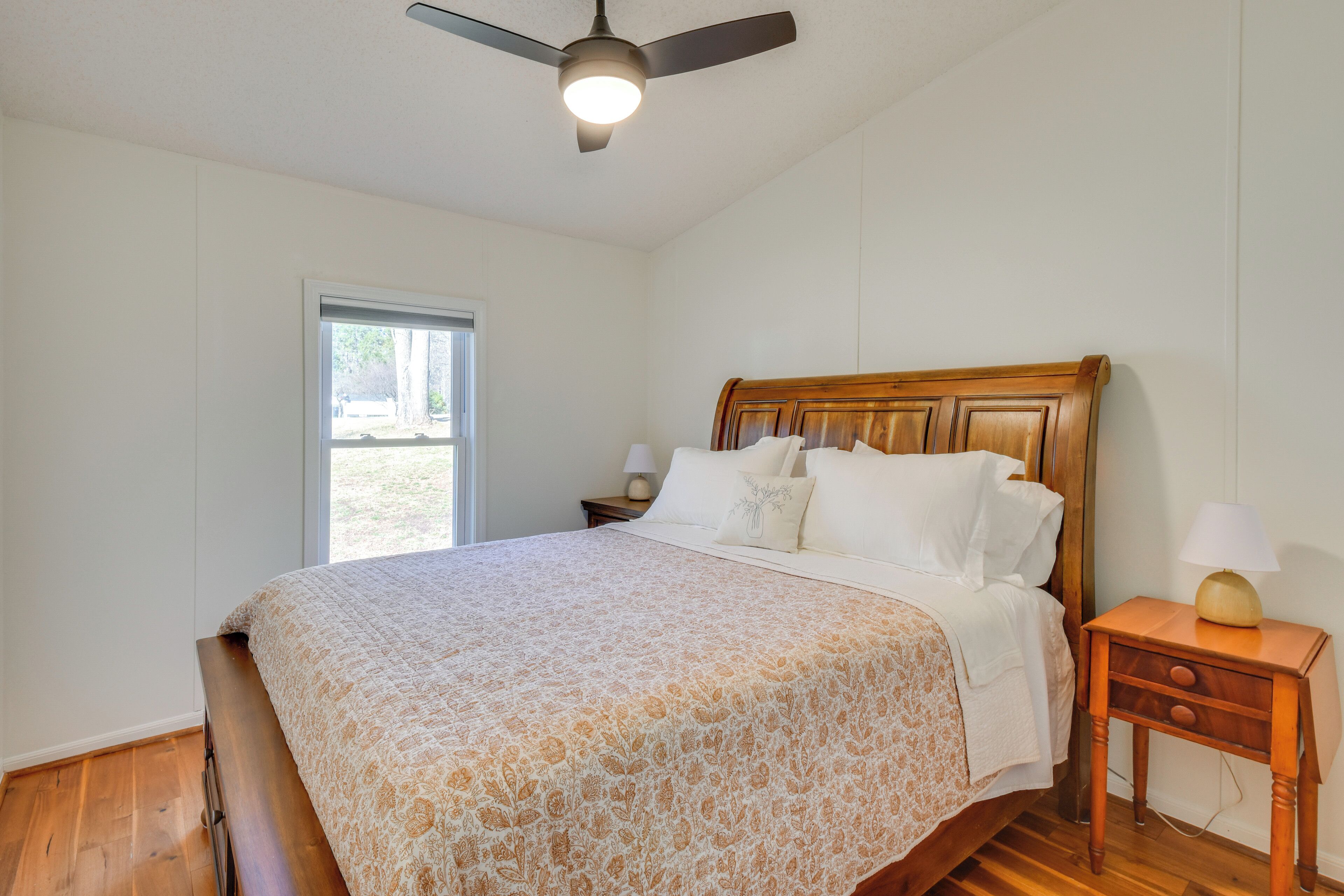 A bedroom with a wooden bed, a nightstand with a lamp, a ceiling fan, and a window.