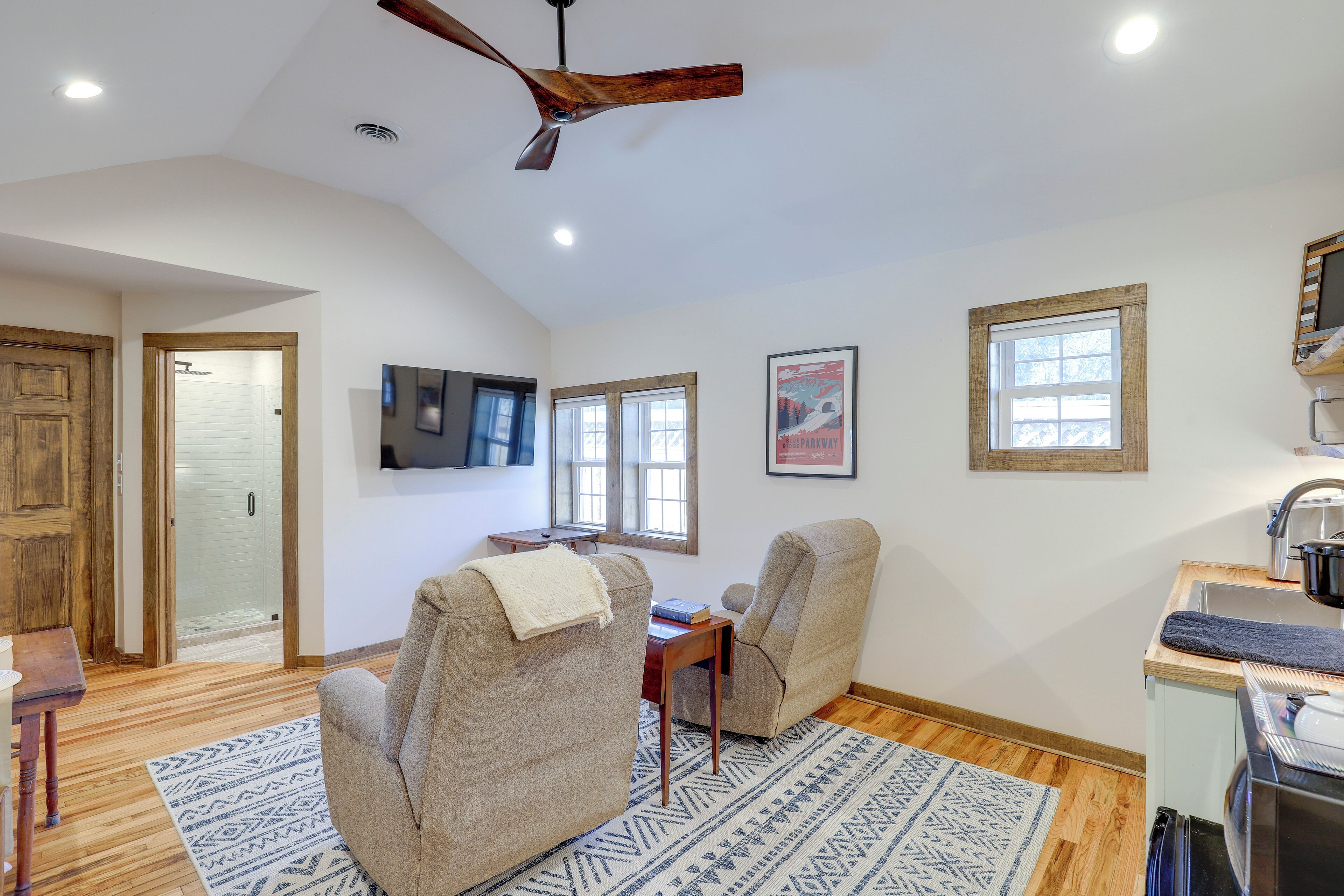 A spacious living room with a high ceiling, a ceiling fan, a wooden door, a television mounted on the wall, and a kitchen area with a sink and countertop.