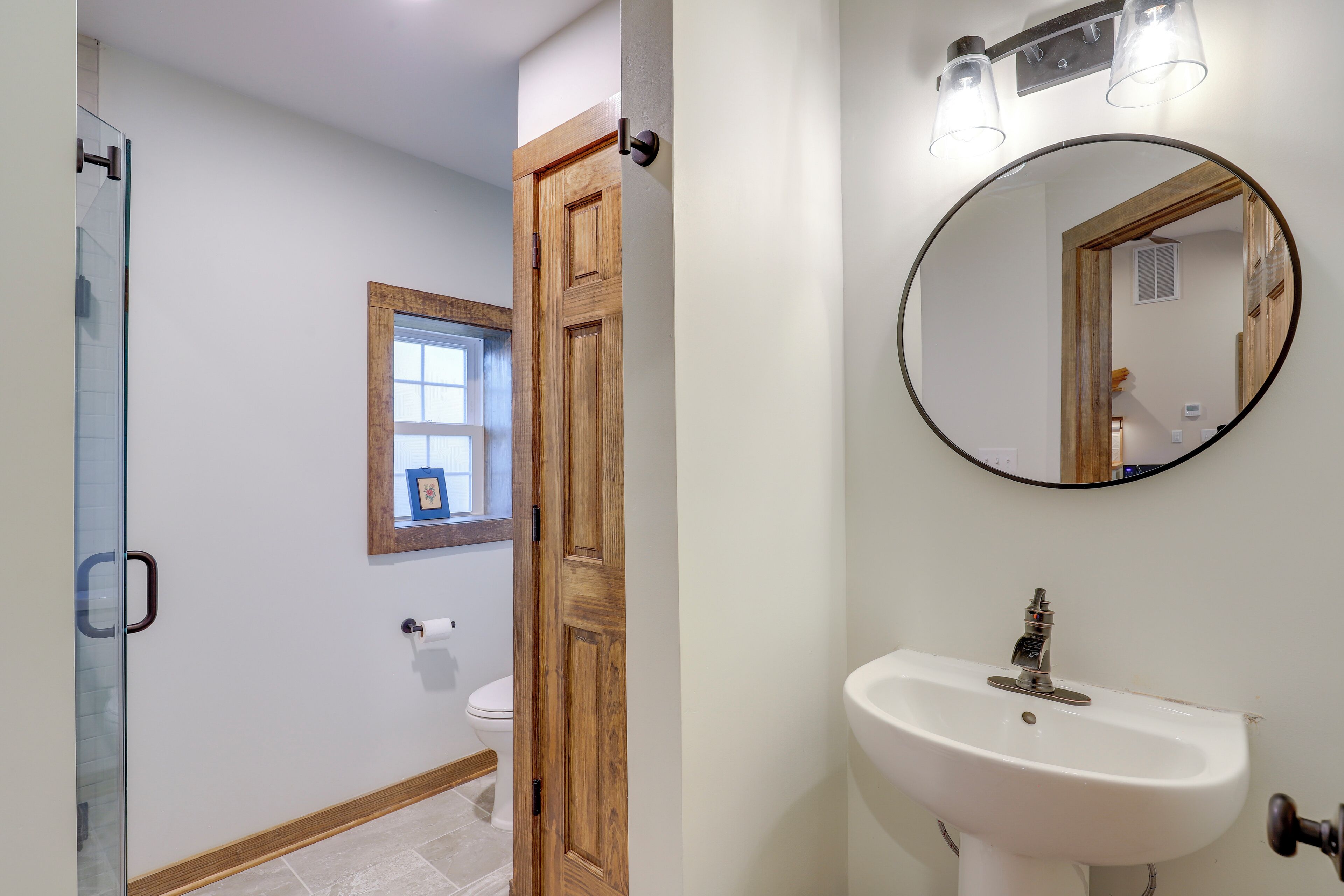 Un baño con puerta de madera, lavamanos blanco, espejo y cabina de ducha de vidrio.