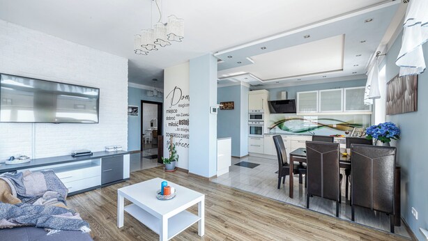A modern kitchen with white cabinets, a dining area with a white table, and a living room with a sofa and a coffee table. - Lion Apartments - The Wave (Gdansk)