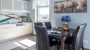 A modern kitchen with white cabinets, a dining table with four chairs, a vase with flowers, and a window with curtains. - Lion Apartments - The Wave (Gdansk)