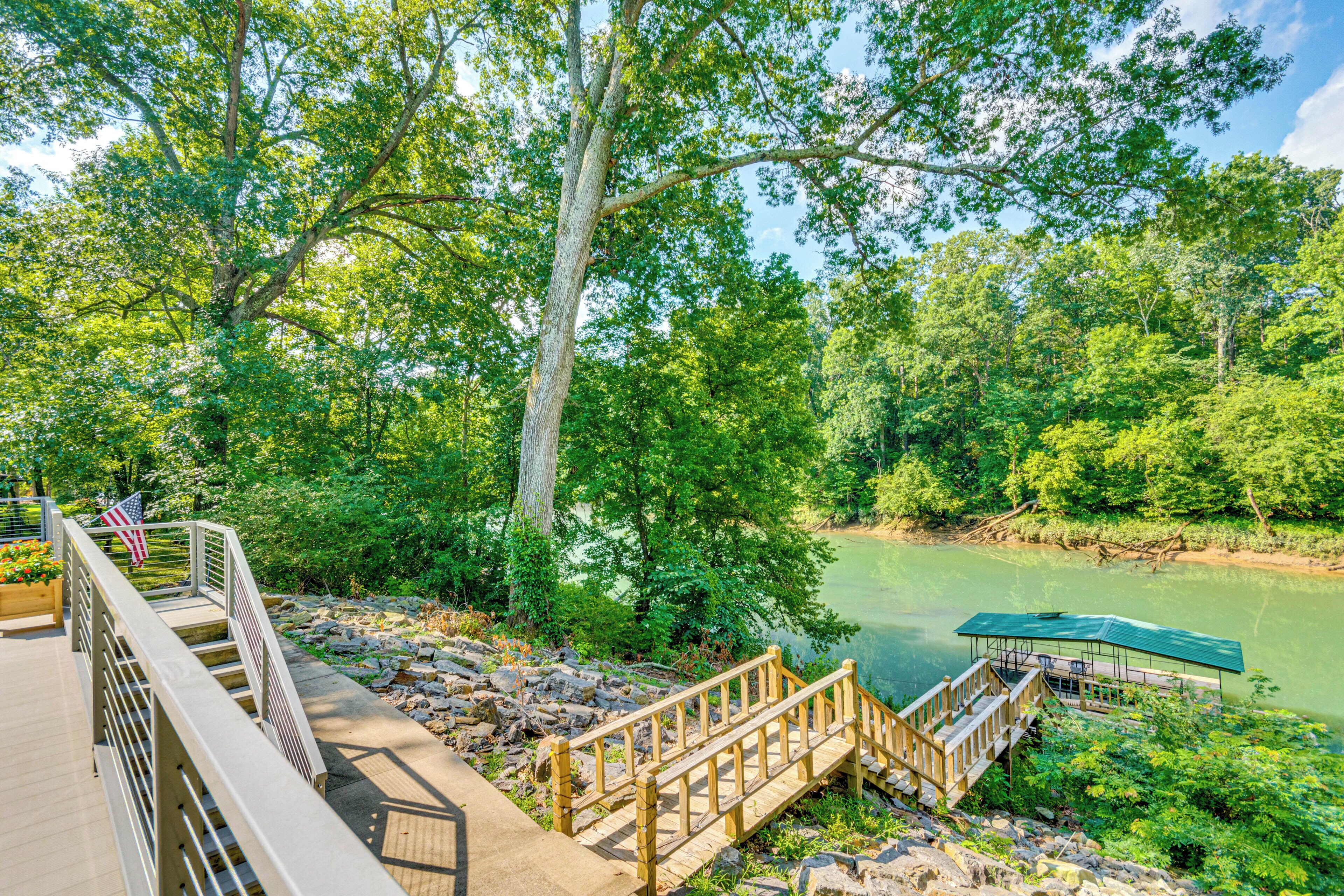 Eine Holzterrasse mit Geländer, eine Anlegestelle, die in einen ruhigen Fluss führt, umgeben von üppiger Vegetation und Bäumen.