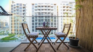 Un balcón con dos sillas y una mesa de madera, una planta en maceta, y vistas a una piscina y edificios altos.