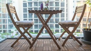 Un balcón con dos sillas de madera y una mesita, una planta en maceta, y vistas a edificios a través de grandes ventanales.