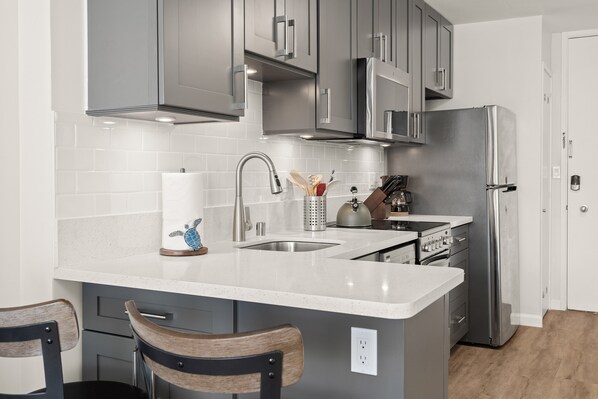 Une cuisine moderne avec des meubles gris, des appareils en acier inoxydable, un plan de travail blanc et deux chaises en bois.
