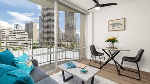 A modern living room with a large window, a ceiling fan, a dining area with two chairs, a table, and a sofa with teal cushions. - Comfy studio in Heart of Waikiki! Quick walk to beach, close to Ala Moana! (Honolulu)
