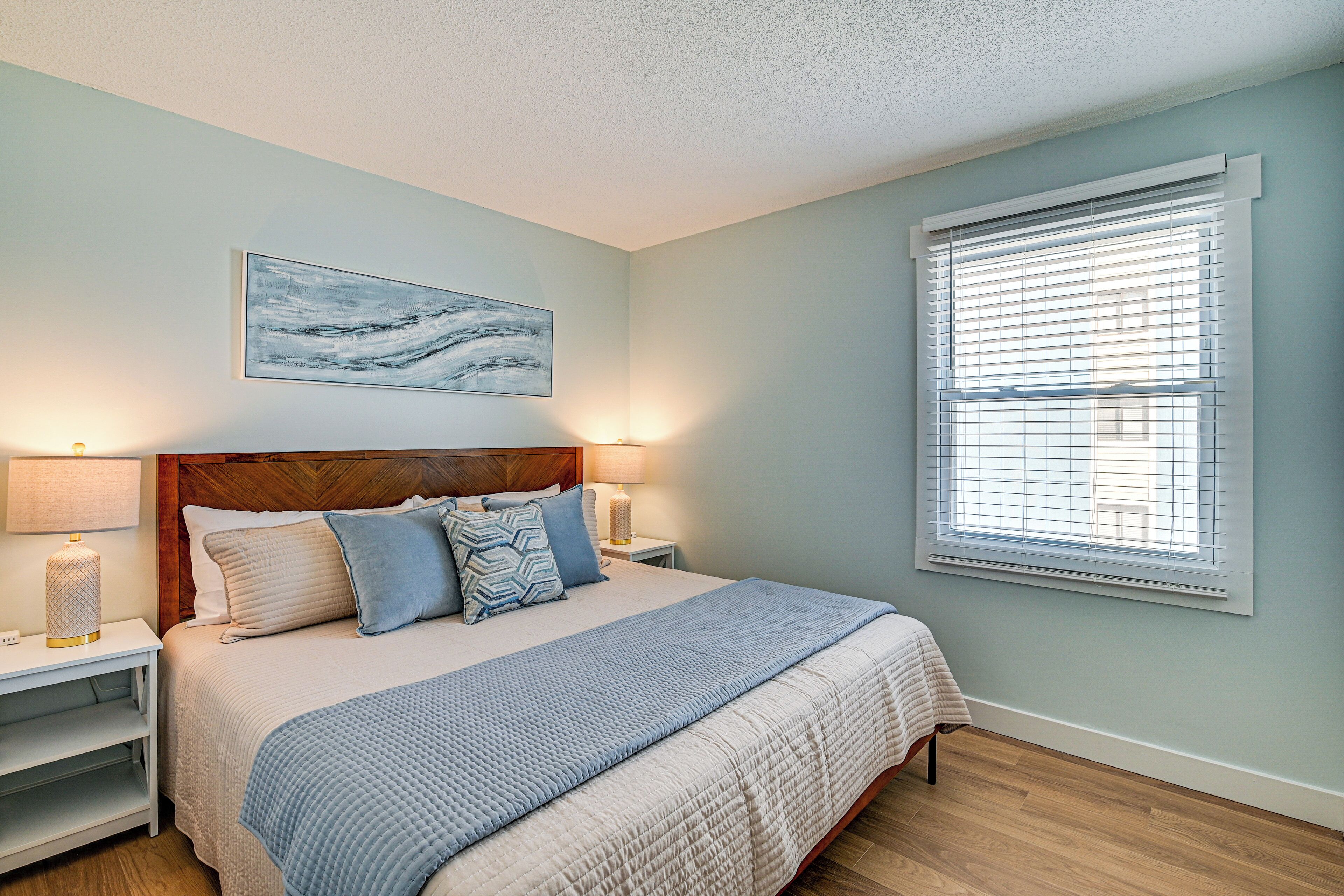 A bedroom with a bed, nightstands, a lamp, a painting, and a window with blinds.