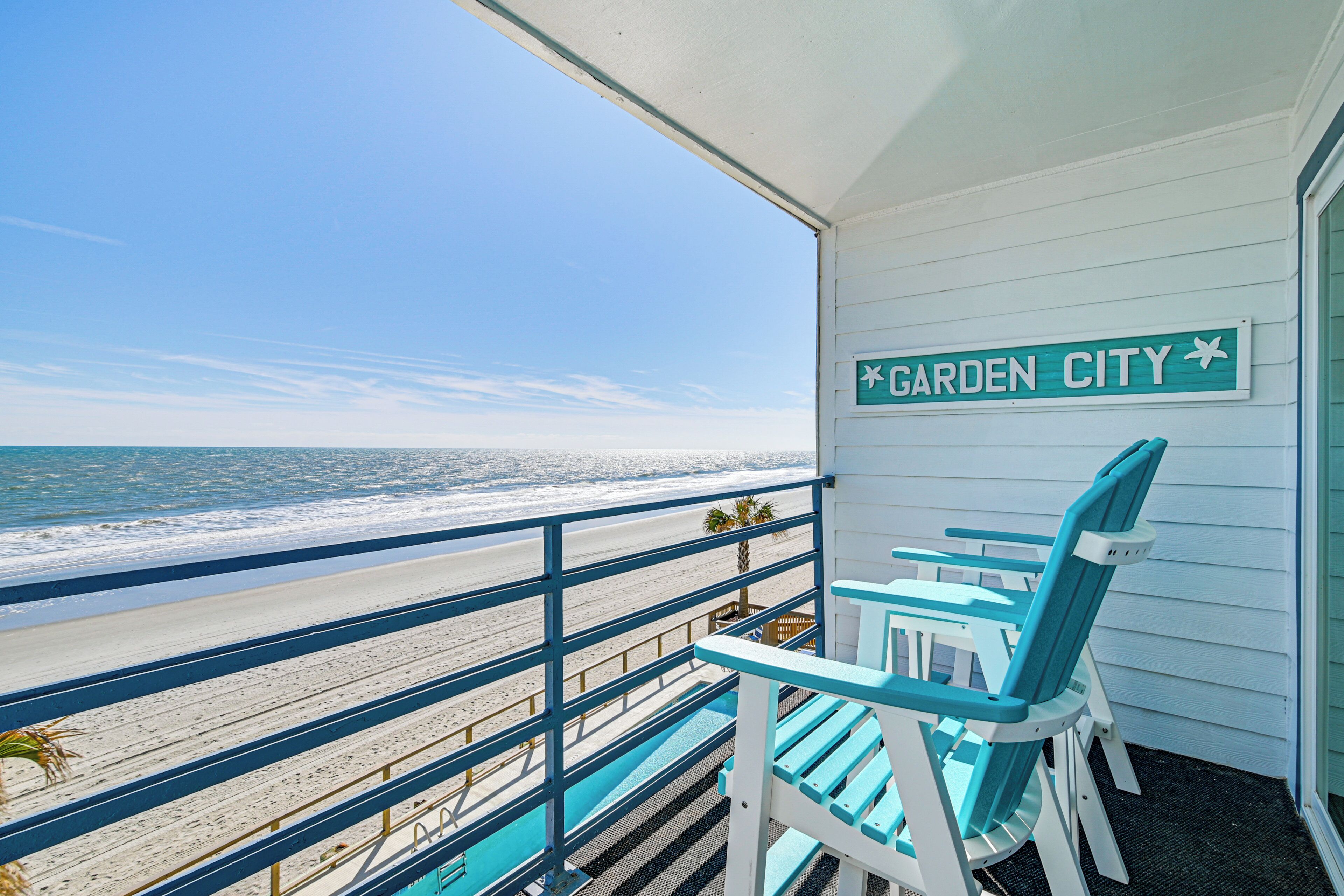 A balcony with a view of the beach and ocean, a sign reading "Garden City," and two blue chairs.