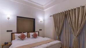 Un lit bien fait, avec du linge de lit blanc et des coussins décoratifs, une table de chevet avec un téléphone et une fenêtre ornée de rideaux beiges.