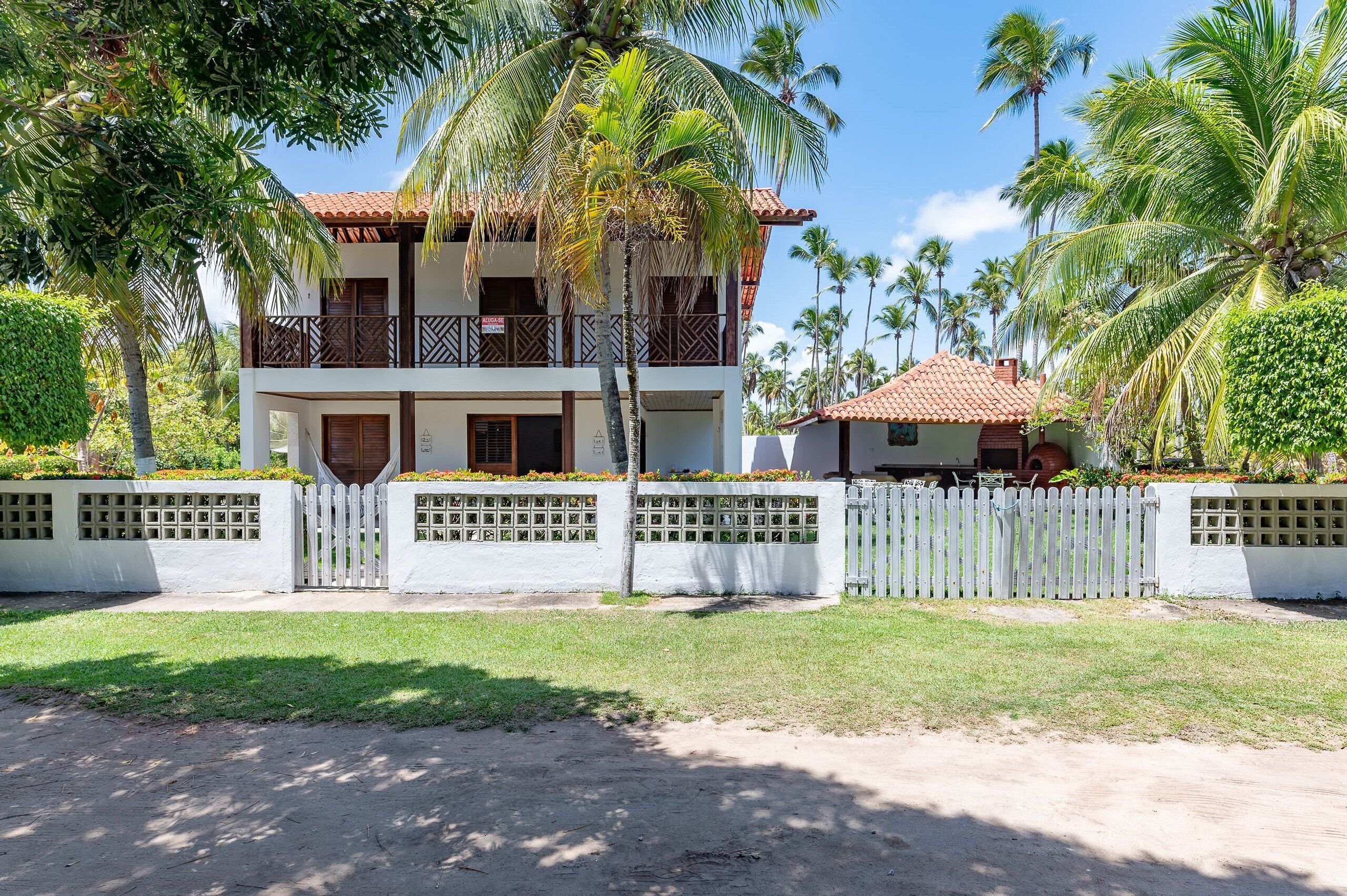 A two-story building with a balcony, surrounded by a white fence and palm trees.