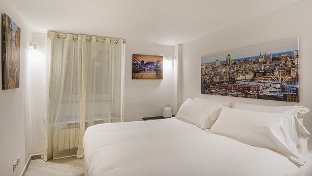 A hotel room with a bed, a nightstand, a lamp, a painting of a cityscape, and a window with sheer curtains. - Galea Studio by Wonderful Italy (Genova)