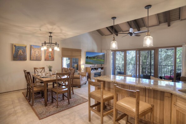 A spacious kitchen with a large island, wooden chairs, and a dining table set for six. There are ceiling fans, pendant lights, and a large window providing a view of the outdoors.