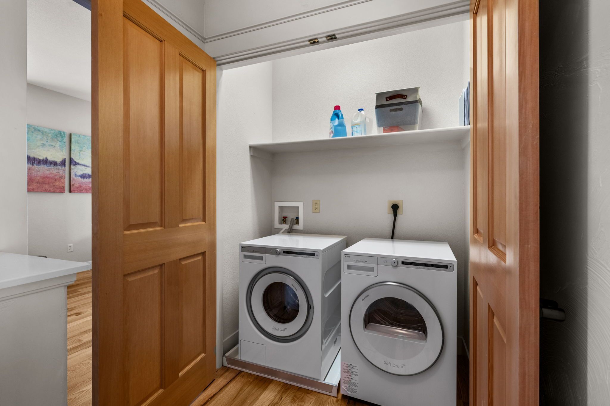 Un cuarto de lavado con dos lavadoras de carga frontal, un estante con productos de limpieza y una puerta de madera.