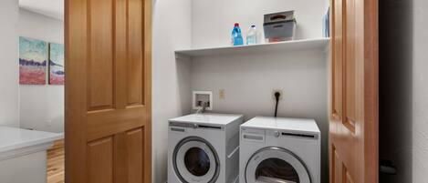 Un cuarto de lavado con dos lavadoras de carga frontal, un estante con productos de limpieza y una puerta de madera.