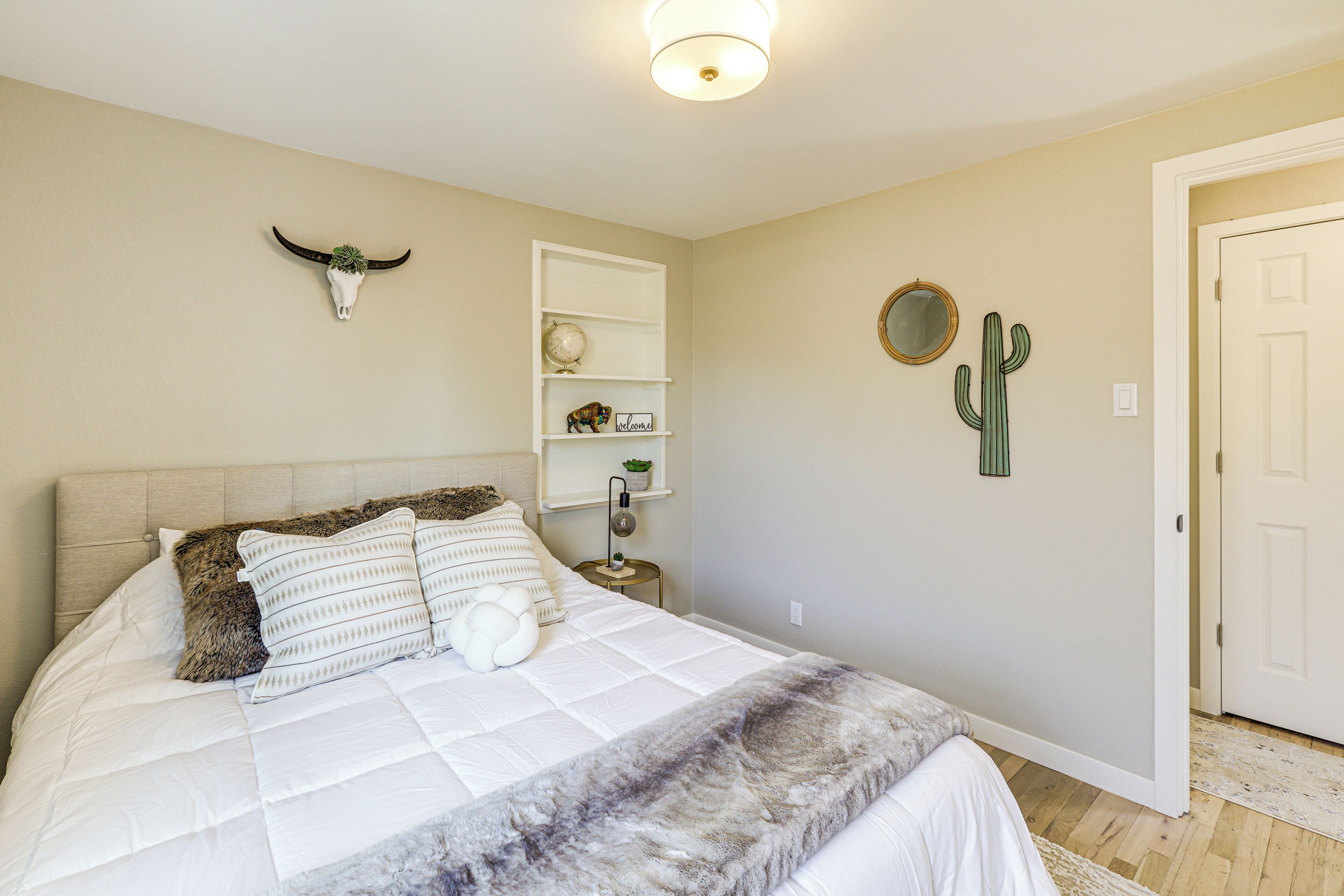 A bedroom with a bed, a nightstand, a wall-mounted shelf, a cactus, and a door.