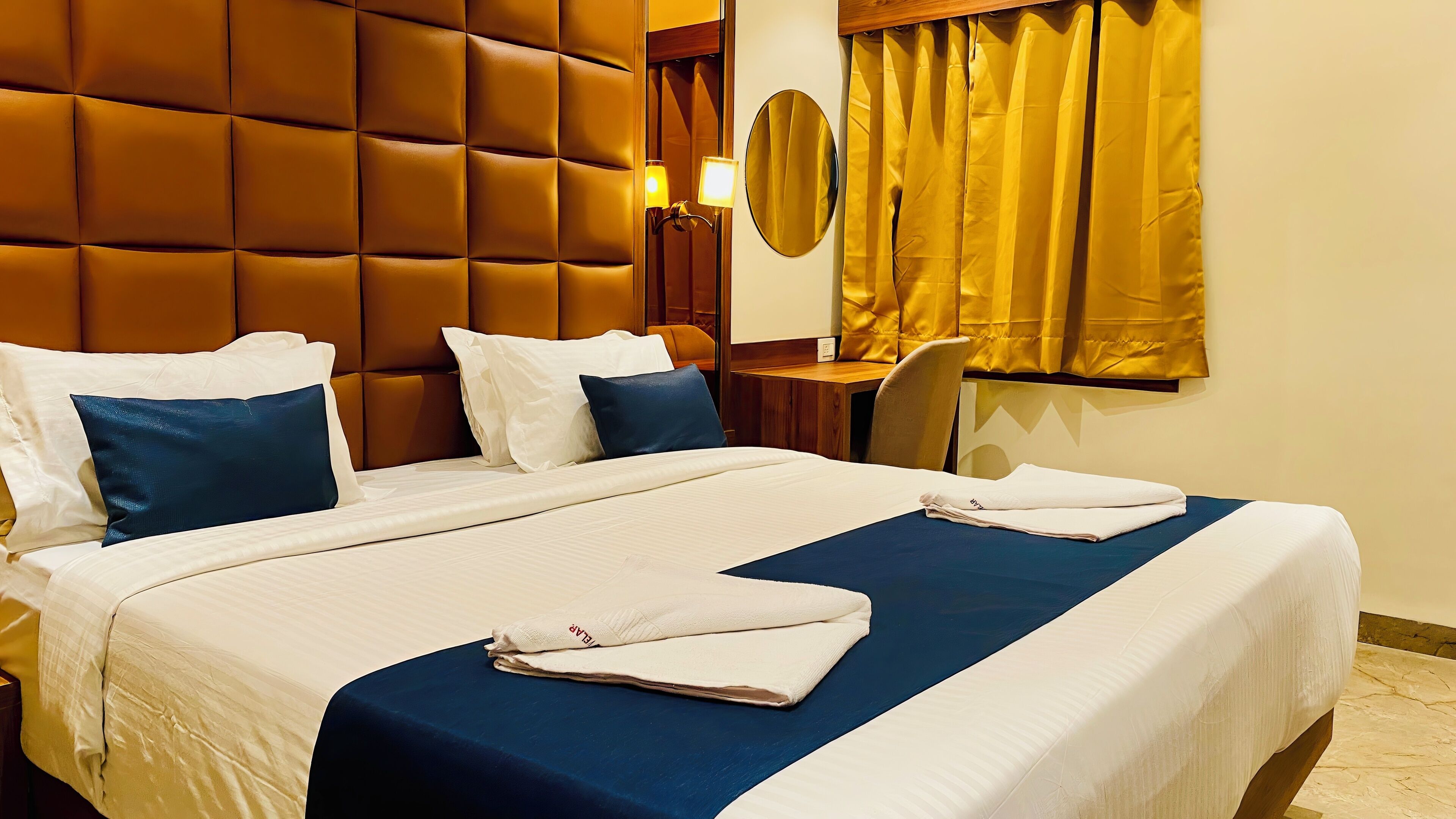 A hotel room with a large bed, two pillows, a bedside table, a chair, a mirror, and a window with curtains.