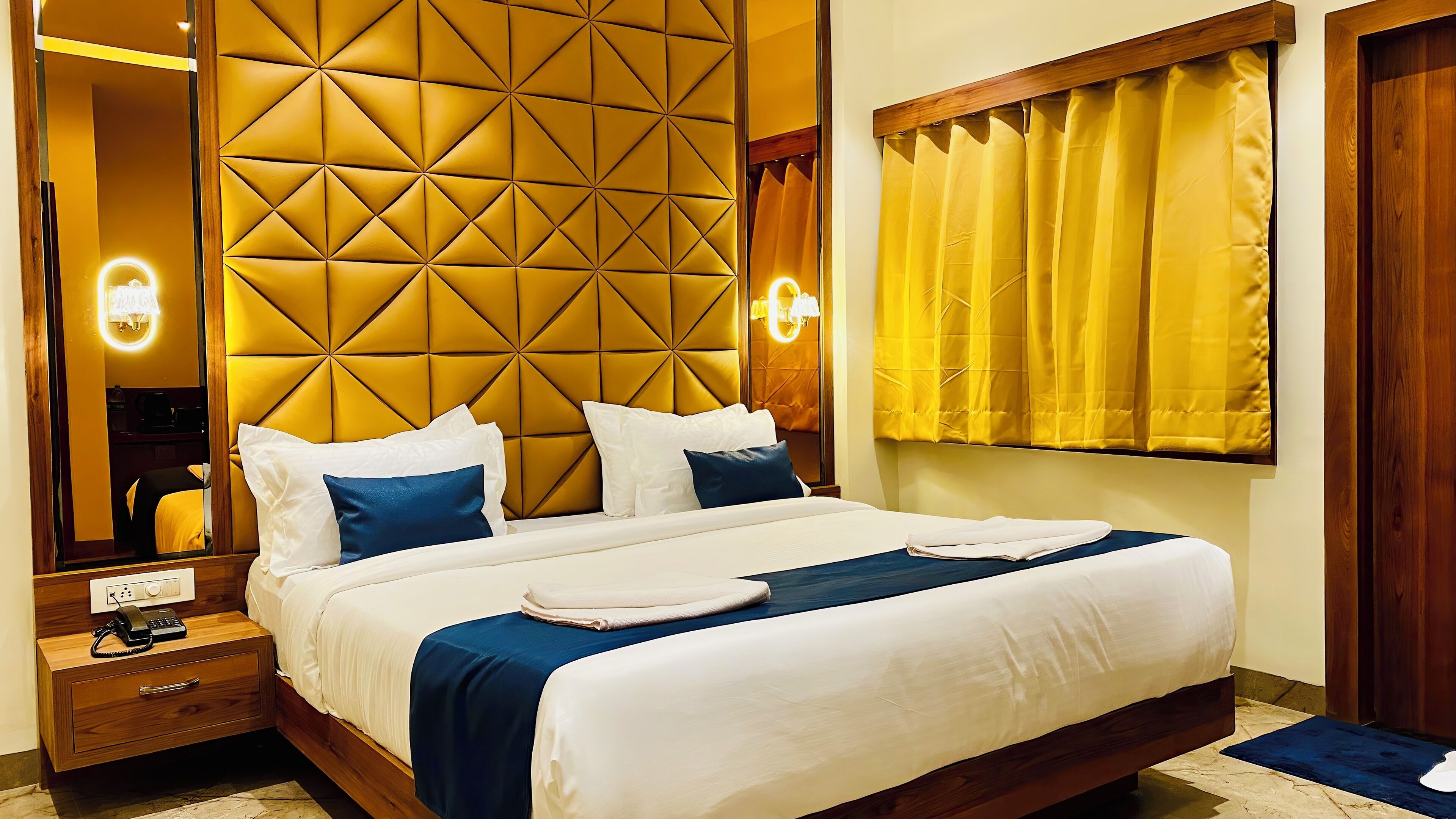A hotel room with a large bed, a wooden nightstand with a phone, a mirror, and a yellow curtain.