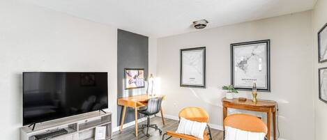 A modern living room with a flat-screen TV, wooden entertainment unit, two chairs, a desk, and framed artwork on the walls.