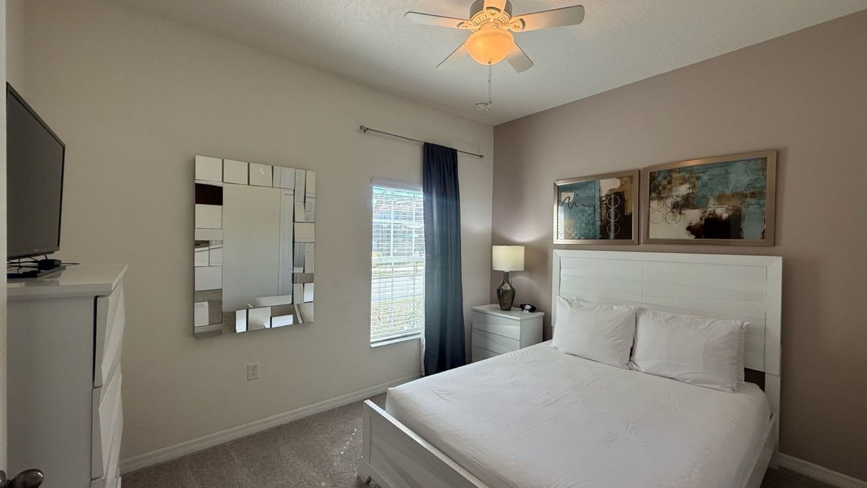 A hotel room with a bed, a nightstand, a mirror, a ceiling fan, and artwork on the wall.