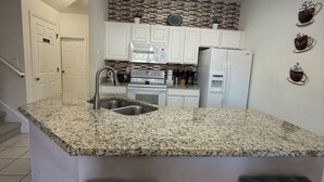 Una cocina con encimeras de granito, armarios blancos, un lavamanos de acero inoxidable, microondas integrado, estufa y refrigerador.