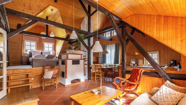 A spacious loft-style room with a high ceiling, wooden beams, a cozy seating area with a sofa and armchairs, a dining table, and a kitchen area. - Loft with sauna and fireplace (Schlitz)