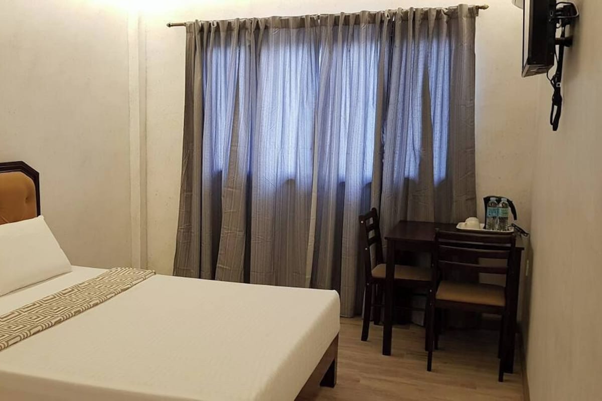 A hotel room with a bed, a desk, and a chair. There is a painting on the wall and a window with curtains.